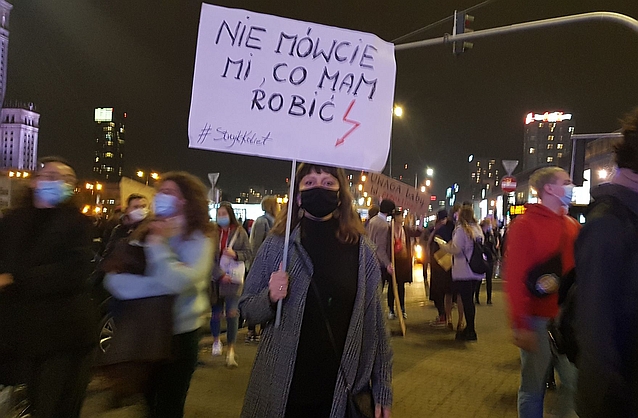 Kobieta stoi w tłumie na ulicy, trzymając transparent z czerwoną błyskawicą i napisem: nie mówcie mi, co mam robić