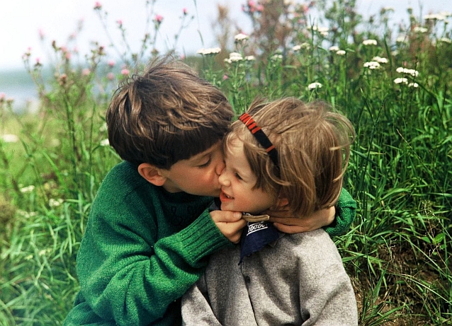 Chłopiec całuje w policzek dziewczynkę, obydwoje siedzą na łące