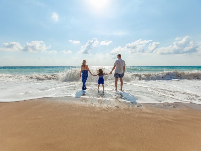 rodzice trzymają córkę za ręce z obu stron stojąc na brzegu morza i plaży
