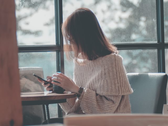 kobieta siedzi bokiem na fotelu przy stoliku przy oknie trzyma rękę na kubku a drugą ręką telefon półdługie włosy zasłaniają jej twarz