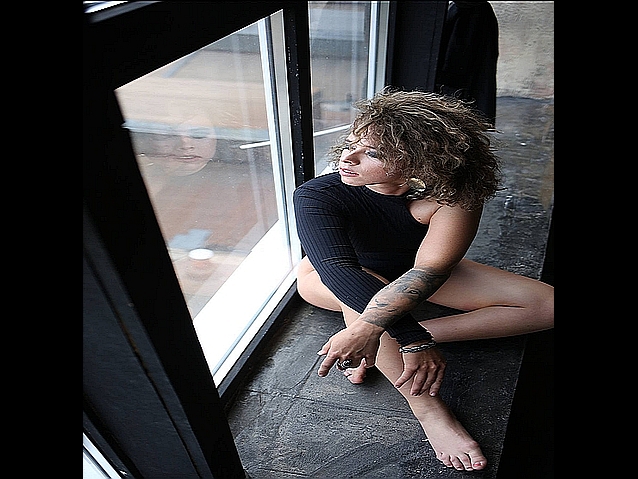 Młoda kobieta siedzi na parapecie patrząc w okno. W szybie odbija się jej twarz