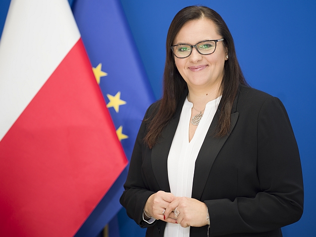 Uśmiechnięta minister Małgorzata Jarosińska-Jedynak pozuje do zdjęcia na tle flag Polsk i Unii Europejskiej