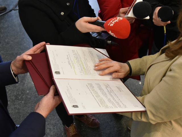 kobieta trzyma w rękach rozłożoną teczkę z pismem naokoło dziennikarze z mikrofonami, nie widać twarzy