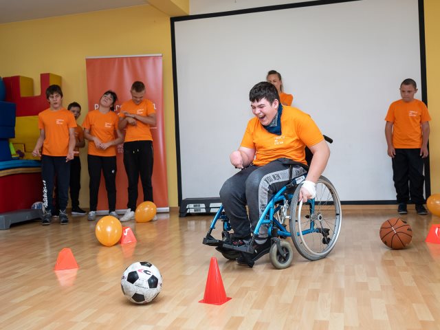 chłopiec na wózku wjeżdża pomiędzy piłki i słupki po bokach stoją inni uczniowe z niepełnosprawnością