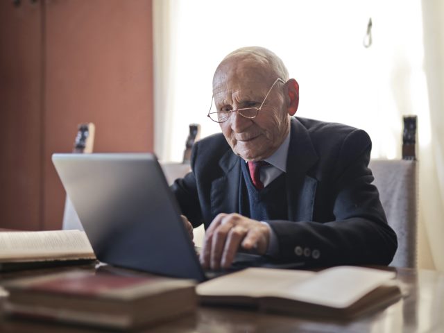 senior mężczyzna w okularach i garniturze siedzi przy biurku przy laptopie na biurku stare książki