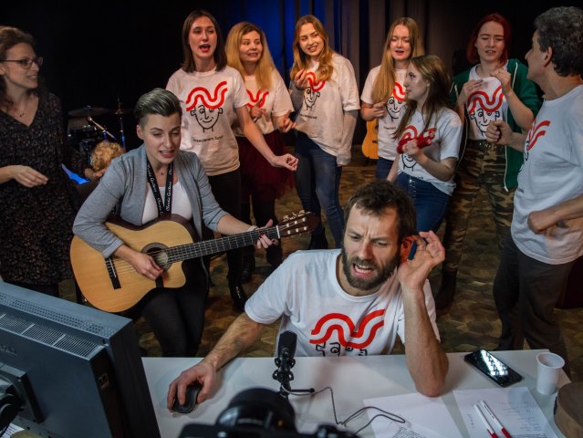 Wolontariusze śpiewają mężczyzna przy mikrofonie za komputerem obok kobieta z gitarą, wokół kilka osób w takich samych koszulkach