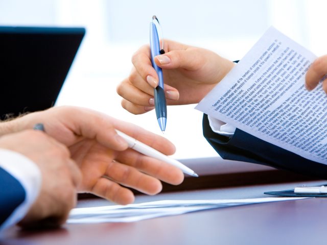 kobiece dłonie trzymają dokument i długopis naprzeciwko męskie dłonie z długopisem przy biurku