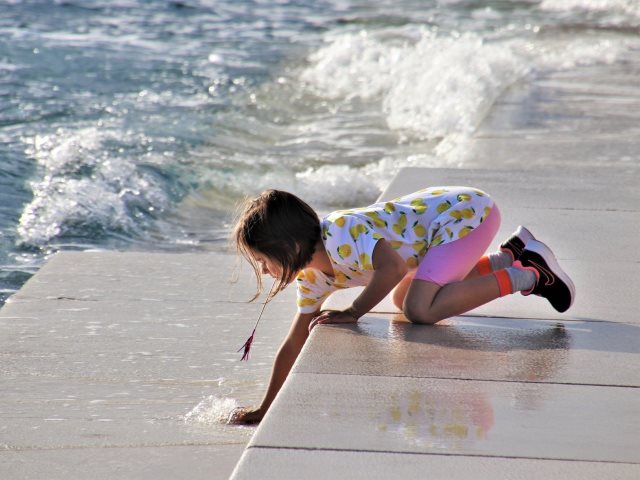 kilkuletnia dziewczynka klęczy na pomoście i schylając się ręką dotyka do morza
