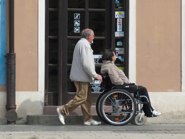 starszy mężczyzna idzie po chodniku pchając wózek z kobietą