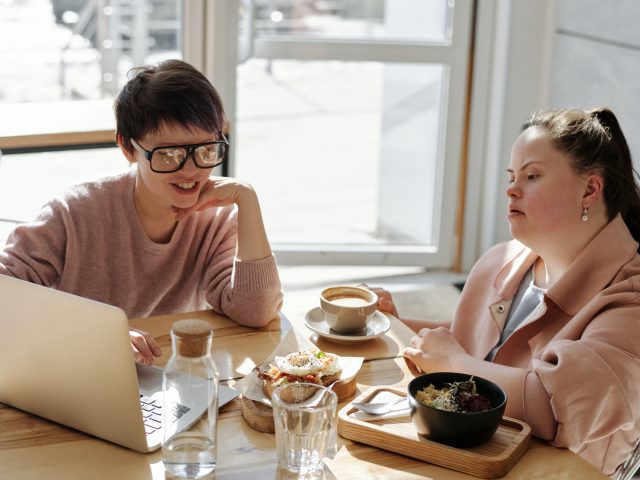 dziewczyna z zespolem downa siedzi przy stole z kawą obok innej dziewczyny patrzącej w laptopa