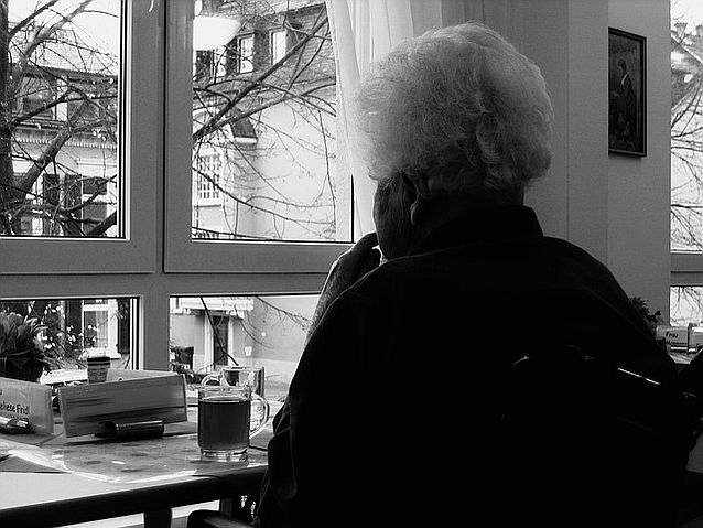 Starsza pani siedzi przy oknie, patrząc w dal. Przed nią stoi szklanka herbaty