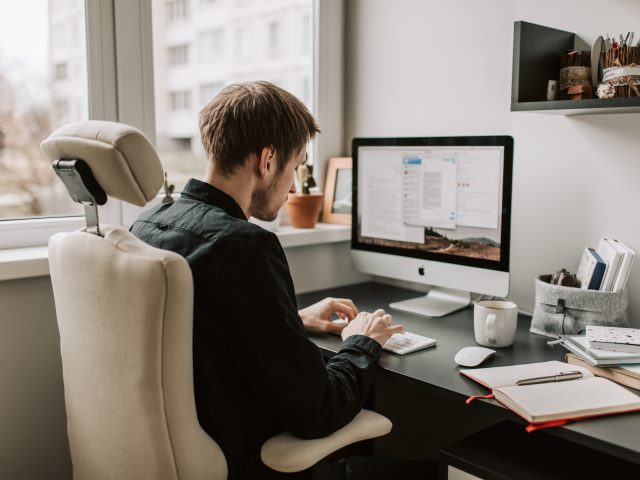 młody mężczyzna siedzi przy biurku i pracuje przy komputerze obok zeszyt i długopis