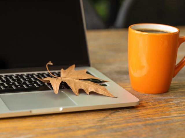 otwarty wyłączony laptop na stole na którym leży jesienny liść obok pomarańczowy kubek
