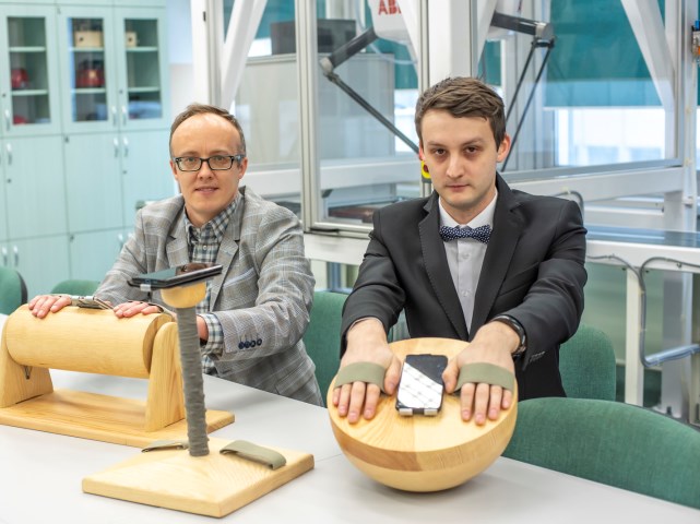 dr inż. Jacek S. Tutak i Paweł Gmiterek siedzą przy stole i trzymają ręce na urządzeniach do rehabilitacji