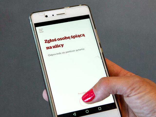 kobieta trzyma telefon w dłoni, wyświetla się napis Zgłoś osobę śpiącą na ulicy