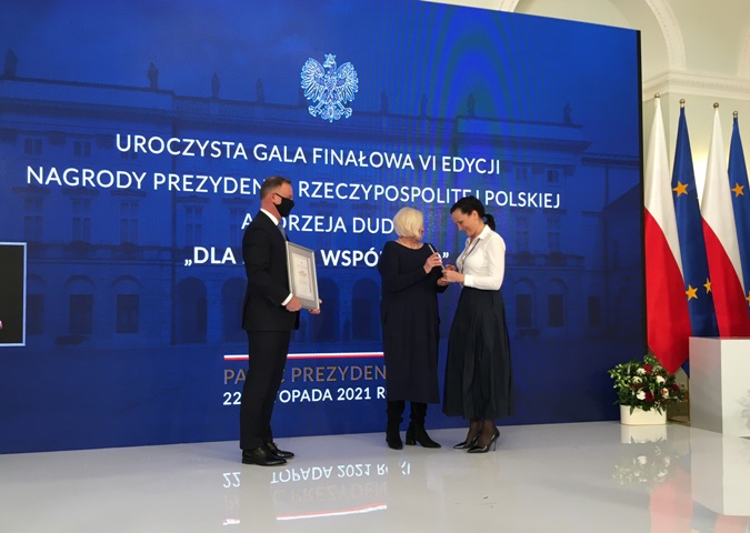 Sala w Pałacu Prezydenckim. Podest. Prezydent Andrzej Duda trzyma dyplom dla Zuzanny Janaszek. Obok Ewa Pawłowska, prezes Integracji przekazuje Zuzannie statuetkę. Zuzanna uśmiecha się.