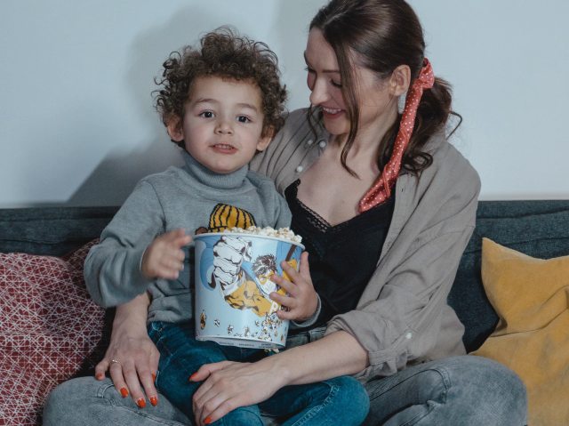 młoda, uśmiechnięta kobieta trzyma na kolanach chłopczyka, ok. 4-5 letniego, która je popcorn z kubełka. Kobieta siedzi na kanapie w domu