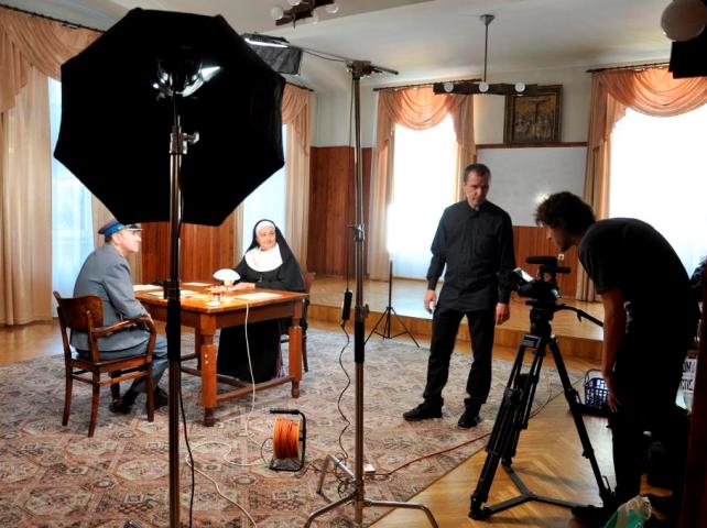 zdjęcie zza kulis z planu filmowego: ksiądz i zakonnica siedzą naprzeciw siebie przy stole w dużym pokoju, obok nich reżyser filmu oraz operator kamery