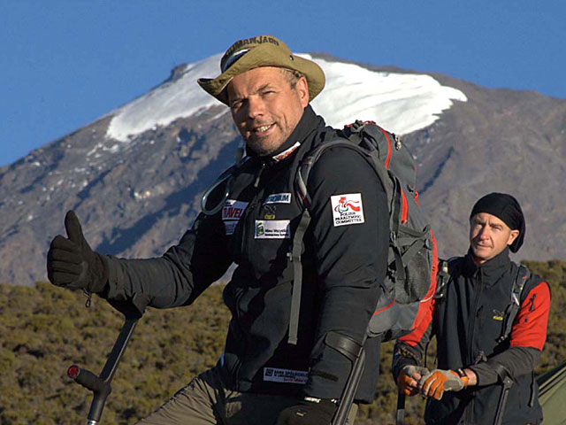 Krzysztof Głombowicz unosi kciuk w górę. Jest ubrany na sportowo z kapeluszem, opiera się na kulach. Jest u stóp Kilimandżaro