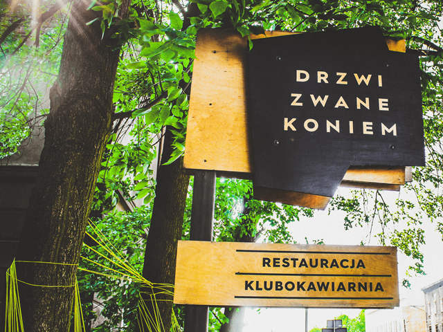 tablica z napisem drzwi zwane koniem oraz restauracja klubokawiarnia wiszą na słupku w tle drzewa
