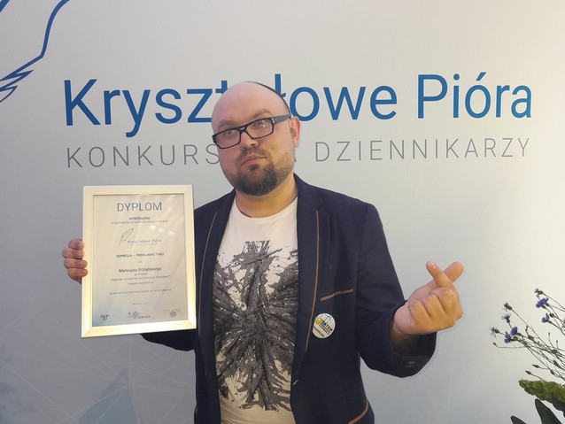 Mateusz Różański pozuje do zdjęcia z dyplomem. Za nim napis: Kryształowe Pióra. Konkurs dla dziennikarzy