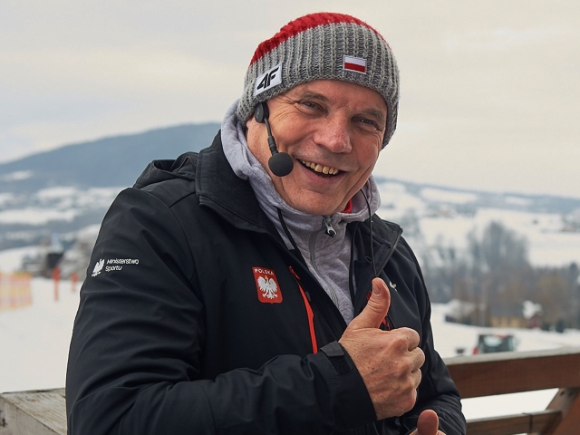 uśmiechnięty Krzysztof Głombowicz pokazuje kciuka w górę. Ma sportową czapkę, bluzę i kurtkę. Ma słuchawkę z mikrofonem przy uchu.. W tle krajobraz zimowy
