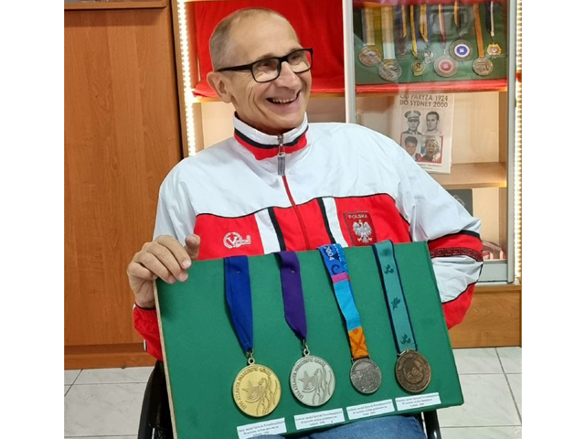 Uśmiechnięty Mirosław Piesak w sportowym stroju na wózku, na kolanach trzyma tablicę ze sportowymi medalami