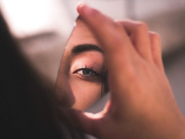 kobieta trzyma w dłoni ułamek stłuczonego lustra, w lustrze odbija się jej oko