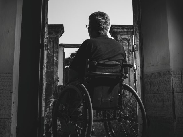 mężczyzna na wózku siedzi tyłem do obiektywu, znajduje się na popękanymi tarasie balkonu w ciemności