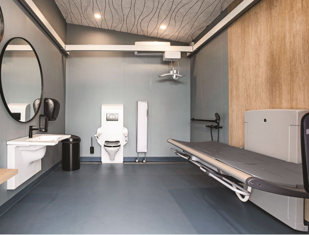 widok na duże pomieszczenie z toaletą. Na wprost jest toaleta, po lewej lustro i umywalka, a po prawej stronie ściany - komfortka, czyli łóżko do przewijania dorosłych osób z niepełnosprawnością