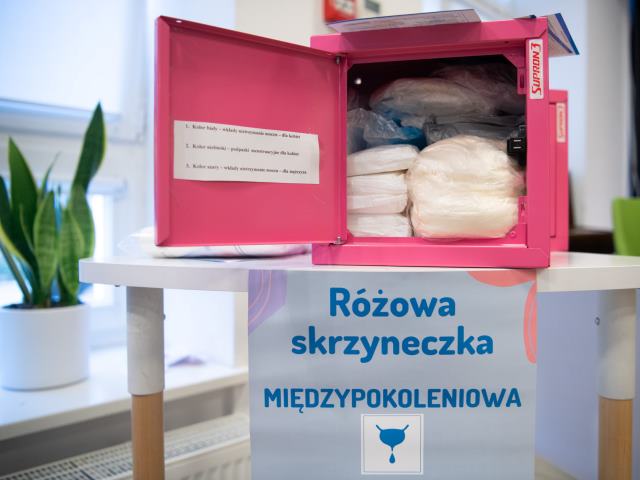 różowa skrzynka z materiałami higienicznymi stoi na białym stoliku oraz napis różowa skrzyneczka międzypokoleniowa