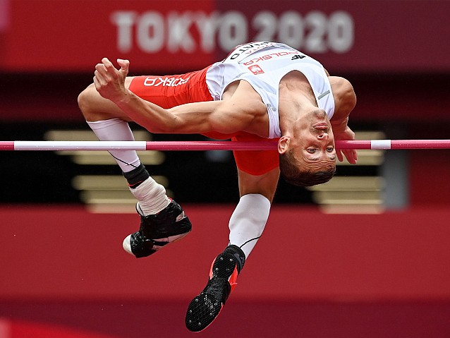 Maciej Lepiato leci nad poprzeczką, odwrócony do niej plecami. W tle napis: Tokyo 2020