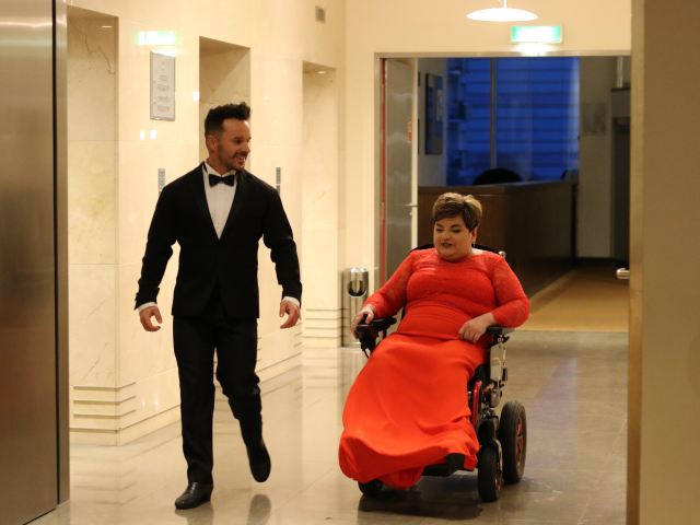 Daniel Qczaj idzie korytarzem z kobietą w czerwonej długiej sukni, poruszającej się na wózku 