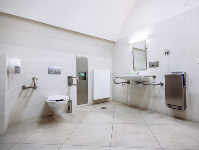 Wnętrze dostosowanej toalety. Na wprost muszla ustępowa z poręczami, po prawej umywalka z poręczami, lustrem, obok niej suszarka do rąk