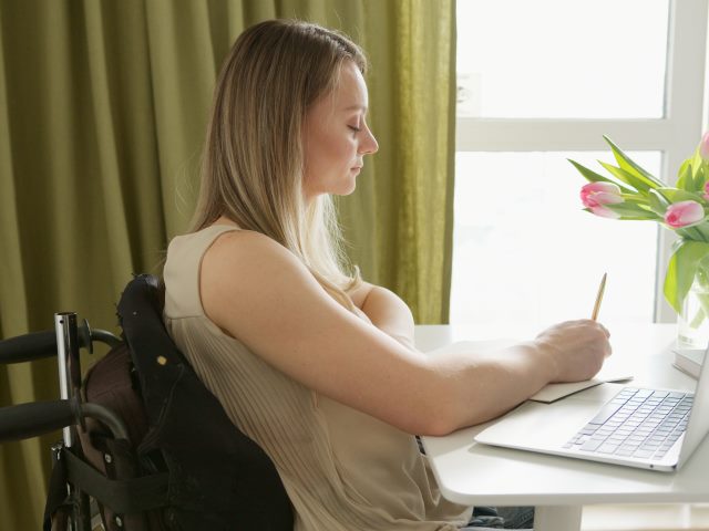 dziewczyna na wózku siedzi przy stole przed otwartym laptopem i coś pisze na kartce