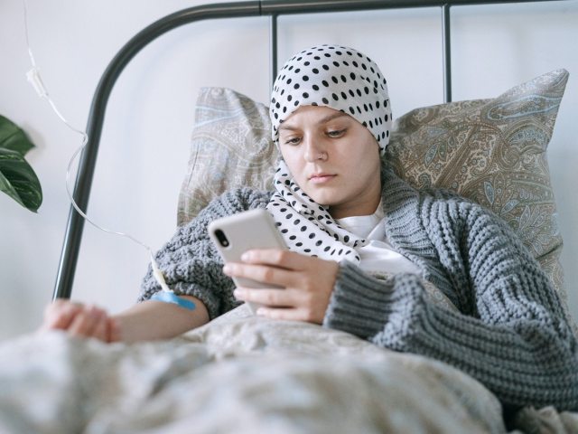 kobieta z chustką na głowie, zakrywającą brak włosów leży w łóżku, przyjmuje kroplówkę, patrzy w telefon