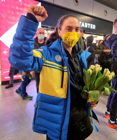 Paraolimpijka w kurtce reprezentacji Ukrainy ma żółtą maseczkę z napisem PEACE, w dłoni trzyma żółte tulipany, drugą rękę unosi na znak walki lub zwycięstwa. Ma uśmiechniete oczy