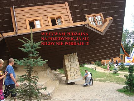 Młody mężczyzna na wózku udaje, że podtrzymuje komin domu naturalnej wielkości, który stoi na dachu. Napis: Wyzywam Pudziana na pojedynek, ja się nigdy nie poddaję!!!