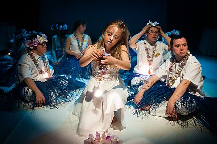 Aktorka tańczy na środku sceny teatralnej, wokół niej siedzą aktorzy. Wszyscy w strojach hawajskich.