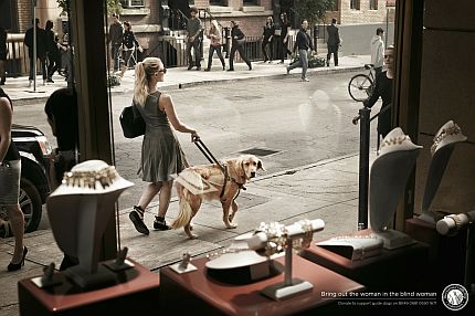 Plakat kampanii. Niewidoma kobieta idzie ulicą, jej pies przewodnik ogląda się za wystawą u jubilera