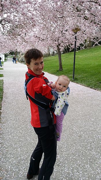 Katarzyna Rogowiec na spacerze z malutką Olimpią w nosidełku. W tle kwitnące drzewa owocowe
