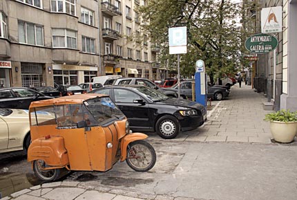 Stary pomarańczowy trzykołowy skuter inwalidzki, w tle nowoczesne samochody