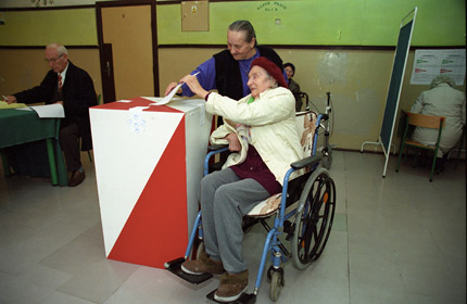 Starsza pani na wózku wrzuca kartę do głosowania do urny. Pomaga jej inna starsza pani