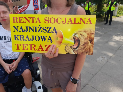 Kobieta trzyma karton z napisem Renta socjalna najniższą krajową teraz i rysunkiem lwa krzyczącego przez megafon. Obok niej idzie młody mężczyzna na wózku. 